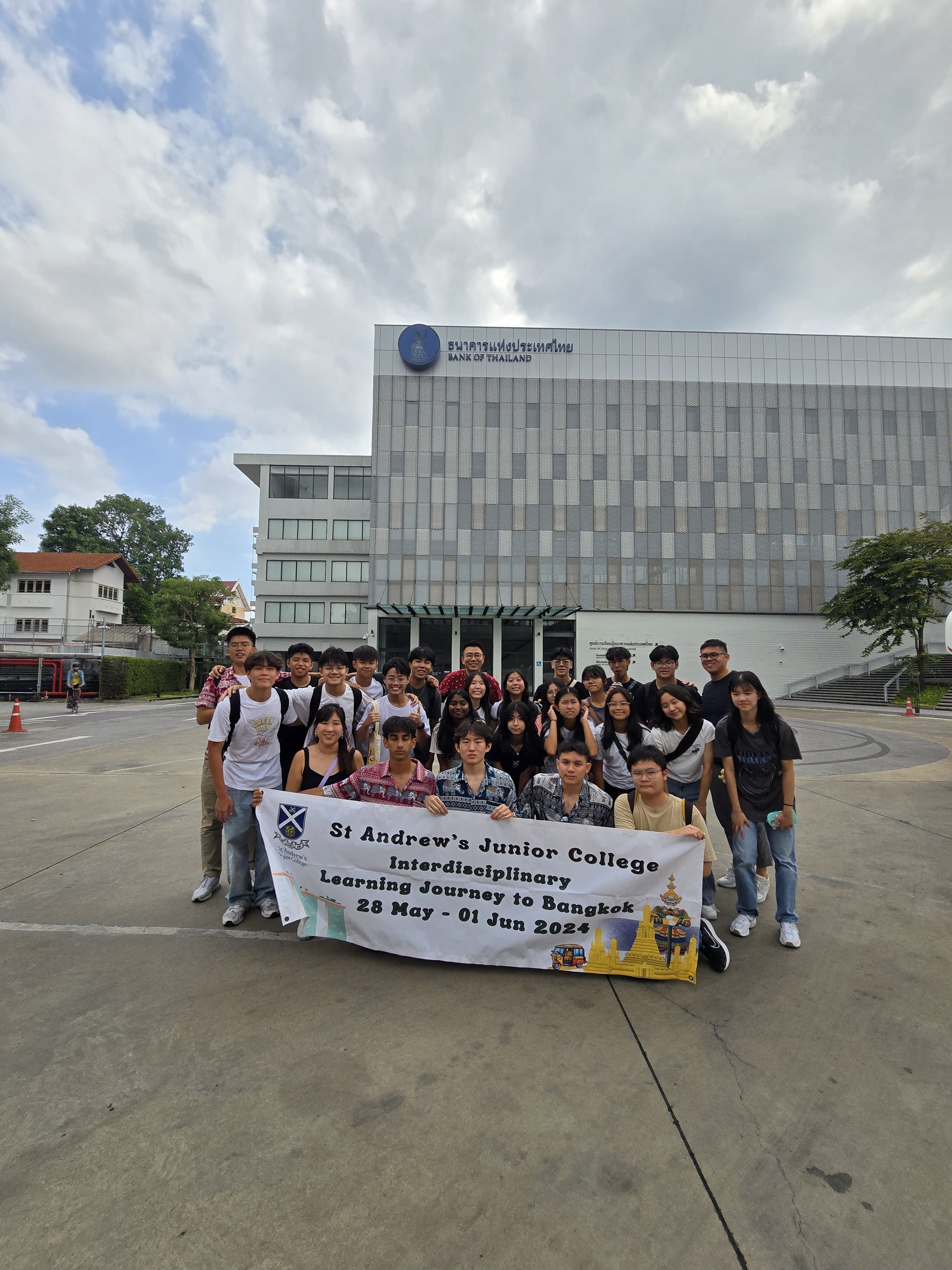 Students from St Andrew's Junior College pose for a group photo outside the Bank of Thailand building, holding a banner that reads 'St Andrew's Junior College Interdisciplinary Learning Journey to Bangkok, 28 May – 01 Jun 2024'.